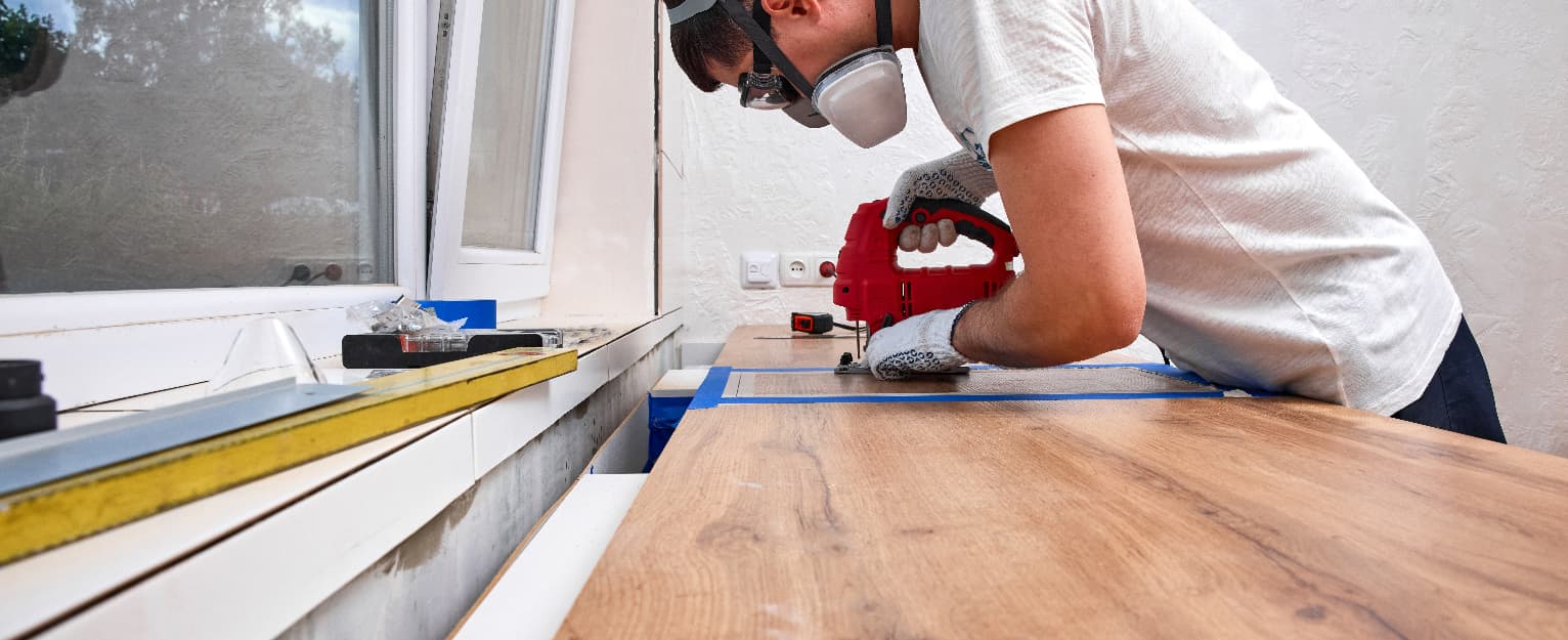 Person wearing safety goggles, gloves and a dust mask carefully cutting a kitchen worktop with a jigsaw, using masking tape and measuring tools to achieve a precise, clean cut during installation.